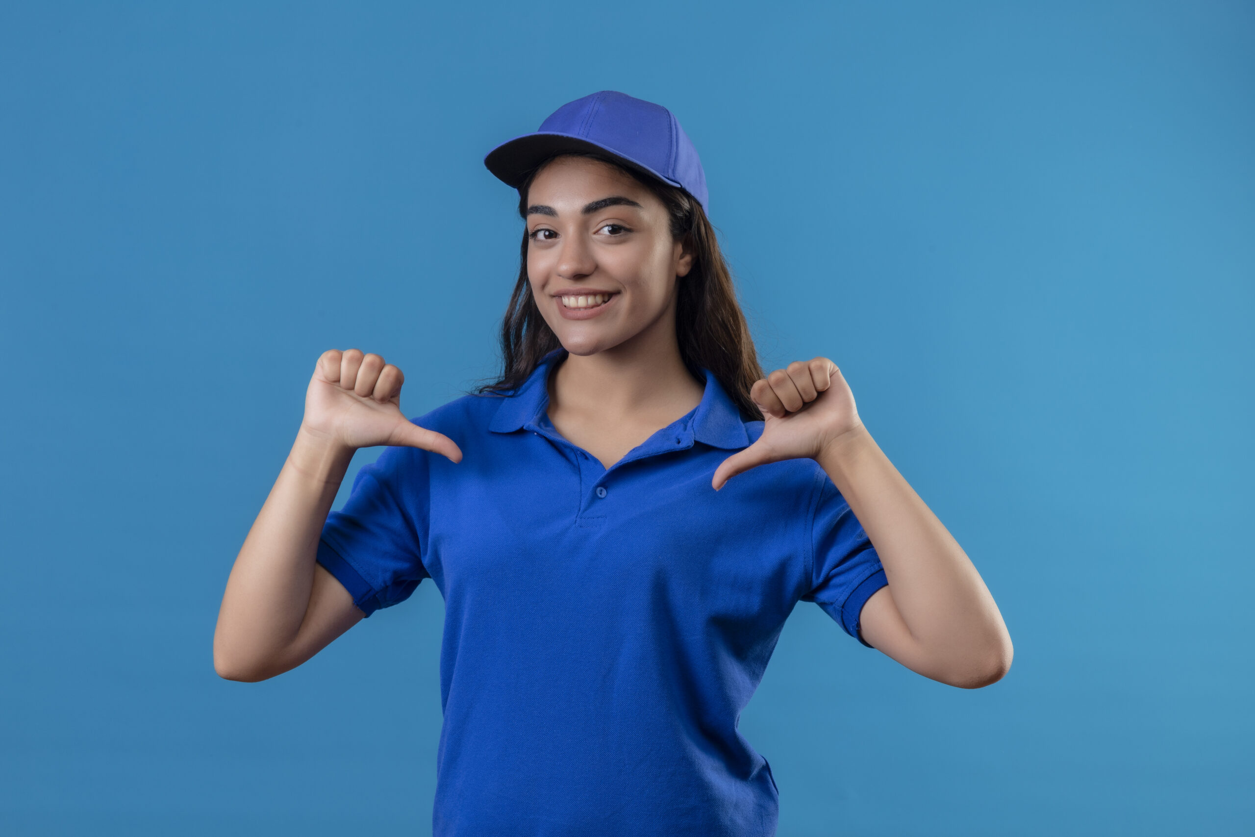young delivery girl in blue uniform and cap looking confident pointing to herself with thumb self-satisfied and proud standing over blue background